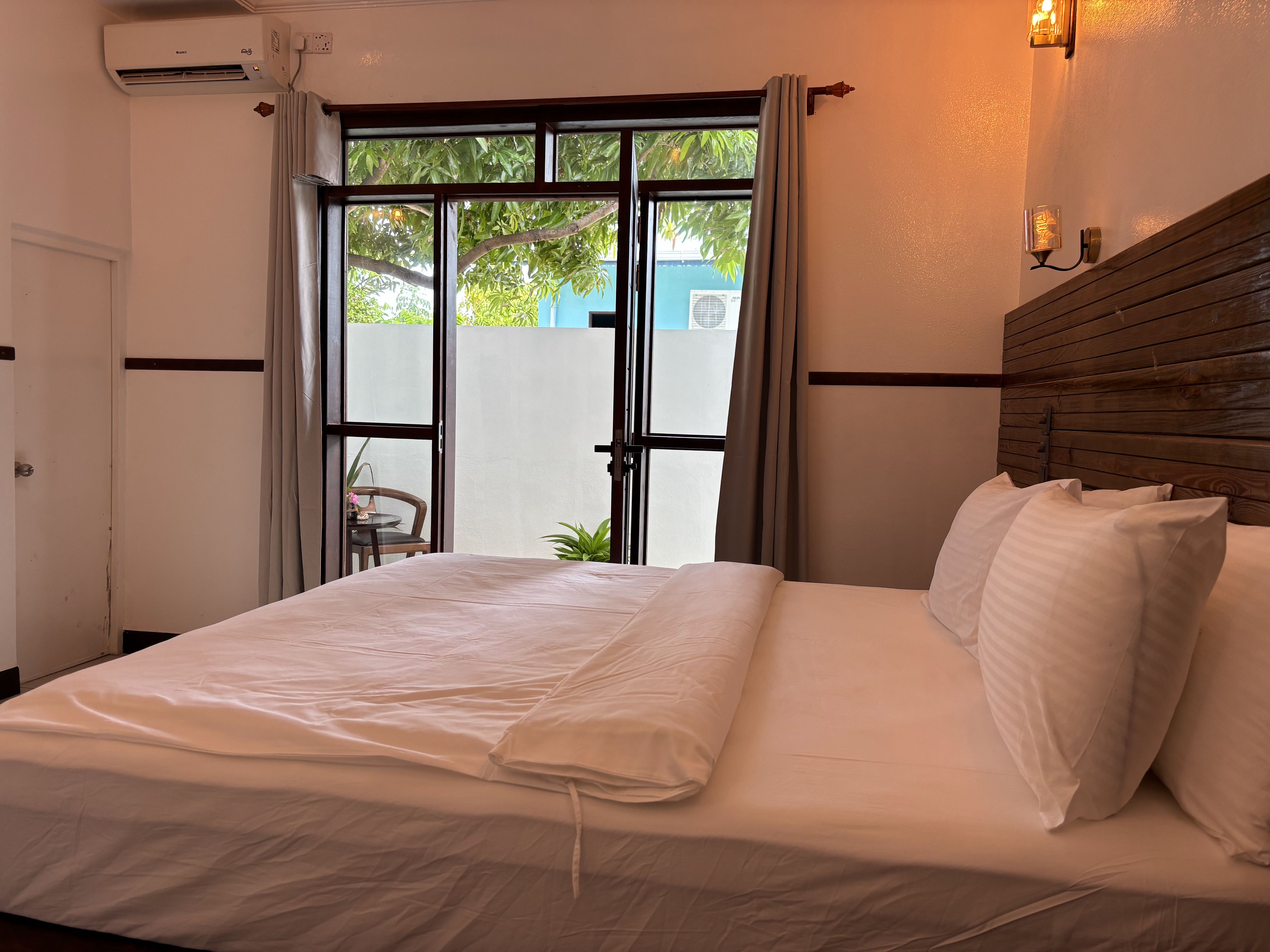 A hotel room with a bed, a window with curtains, a wall-mounted light fixture, and a view of greenery outside.
