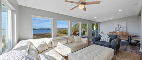 A spacious living area with a large sectional sofa, a smaller sofa, and a coffee table. There is a ceiling fan, large windows offering a view of the outdoors, and a painting on the wall.