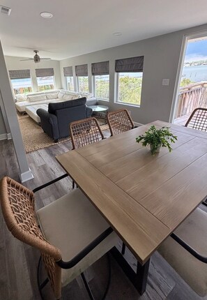 A dining area with a wooden table and wicker chairs, a gray sofa, and a view of a body of water through large windows. - Bay-view 5BR Gulf Shores Home, Sleeps 12, and Easy Beach Access (Gulf Shores)