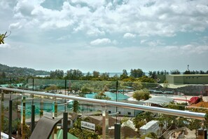 A view from a balcony with a railing, overlooking a parking area, a building with a green roof, and a body of water in the distance. - Kamala - Peaceful Jungle View (Tambon Kamala)
