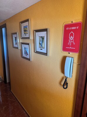 Un couloir orné de tableaux encadrés et sur lequel est affiché un panneau rouge indiquant les numéros de téléphone d’urgence.