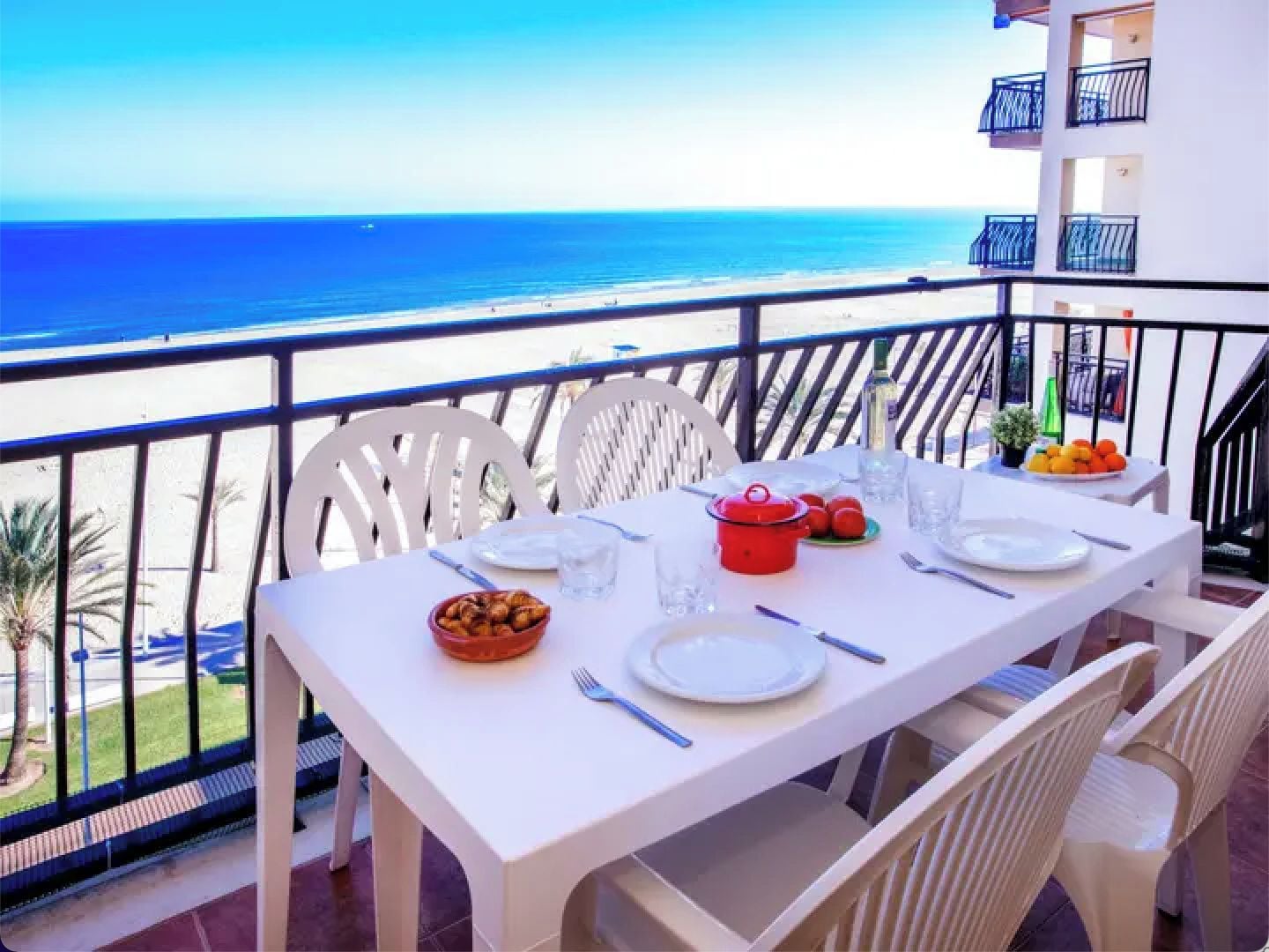 A balcony table set with plates, cutlery, a red pot, and a bowl of fruit, overlooking a beach.
