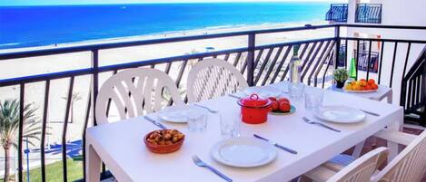 A balcony table set with plates, cutlery, a red pot, and a bowl of fruit, overlooking a beach.