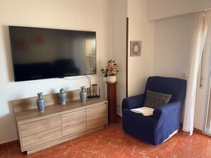 A living room with a flat-screen TV, a blue armchair, a wooden console table with decorative vases, and a potted plant. - Gandia Beach, First-Line Beachfront (Grau i Platja)