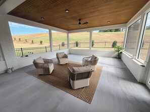 Une terrasse couverte, aménagée avec du mobilier d’extérieur, de grandes fenêtres et une vue sur un espace paysager.