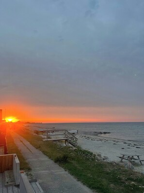 Ein Strand mit einer Holzpromenade, die zum Wasser führt, Picknicktischen und einem Sonnenuntergang im Hintergrund.