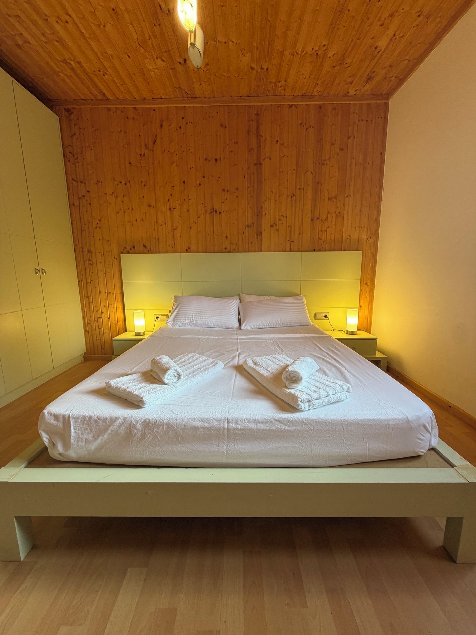 A single-bed room with wooden panel walls, a white bedspread, and bedside lamps.