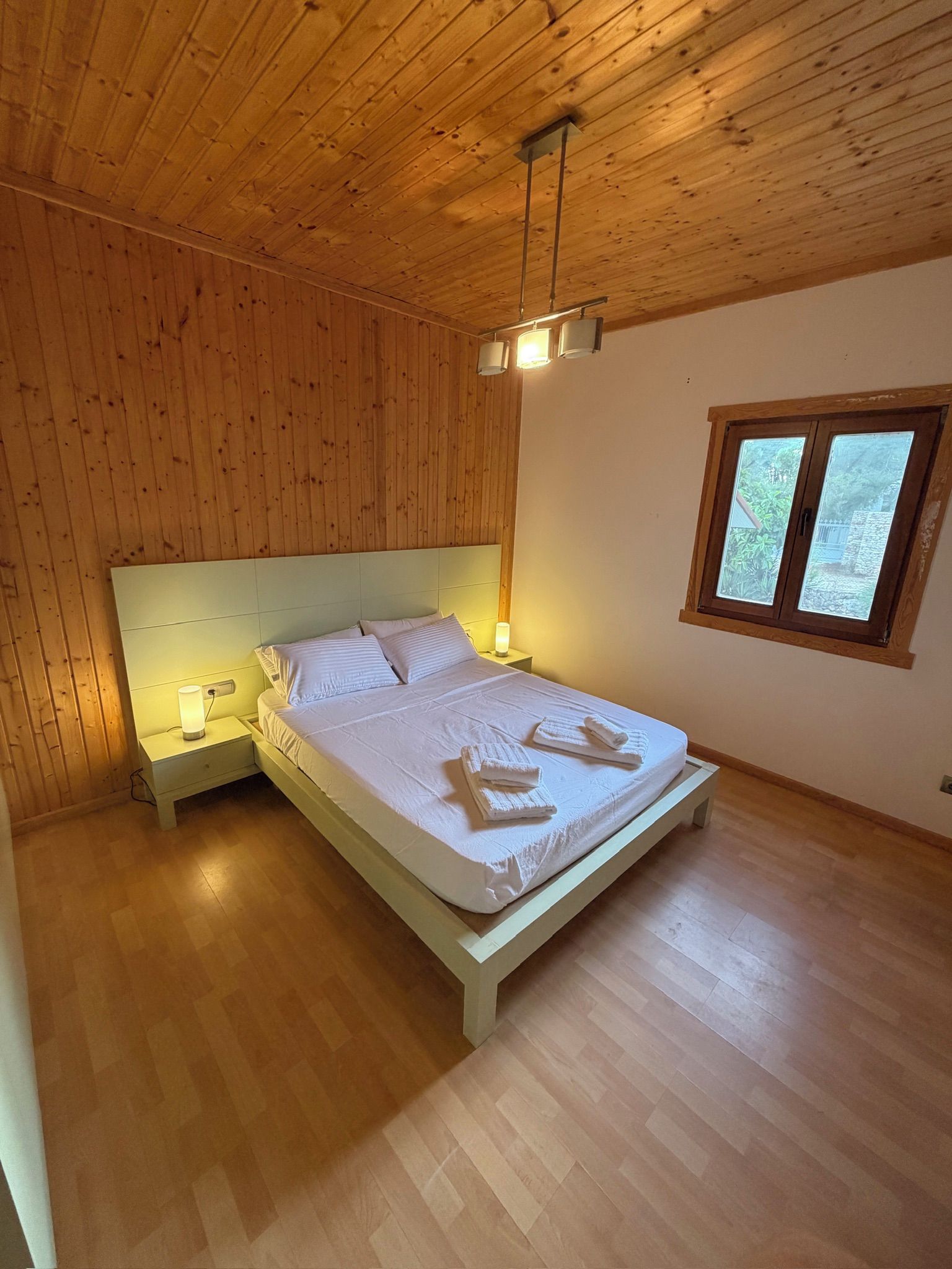 A bedroom with a wooden ceiling, a bed with white linens, a nightstand, and a window with a view of greenery.