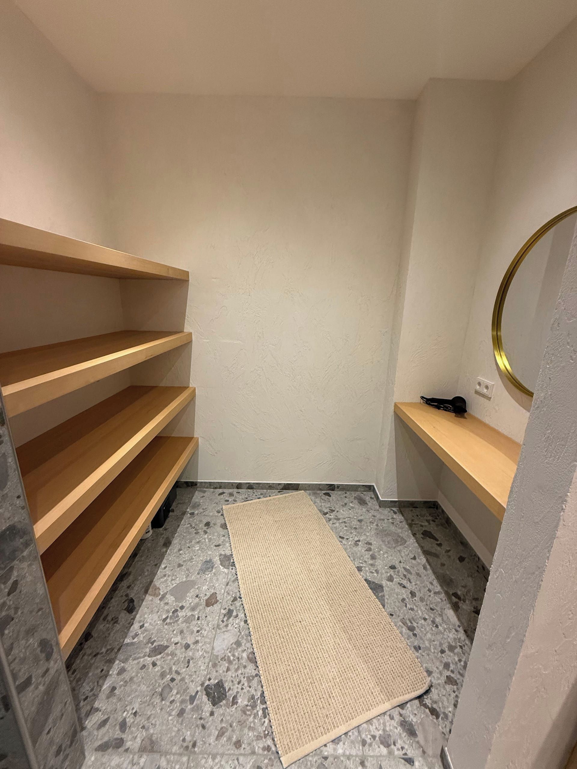 Een kleine, lege kamer met houten planken, een spiegel en een vloerkleed op een geruite vloer.