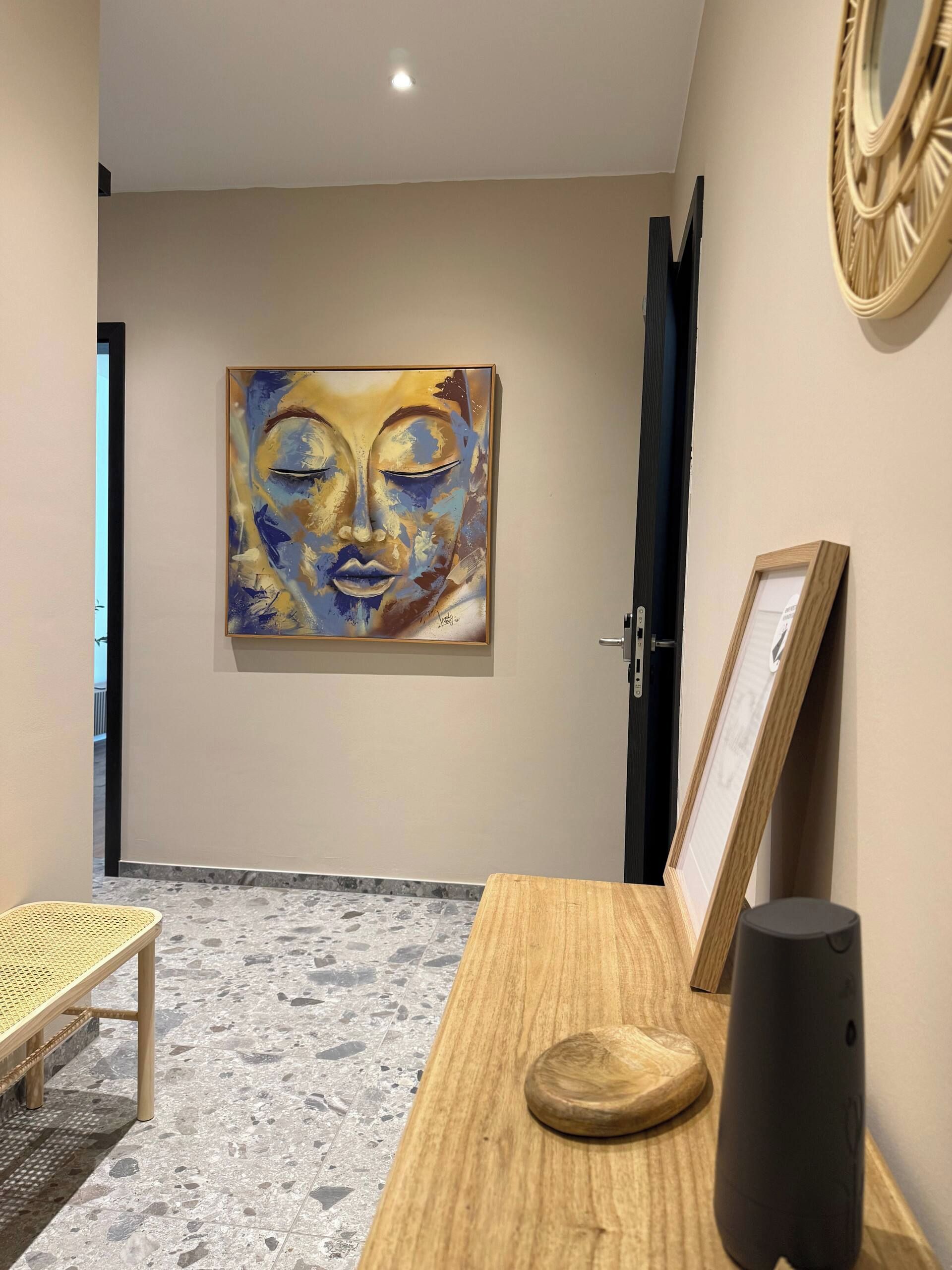Un couloir orné d’un grand tableau abstrait au mur, avec un banc en bois, une petite table sur laquelle est posé un vase, et une porte.