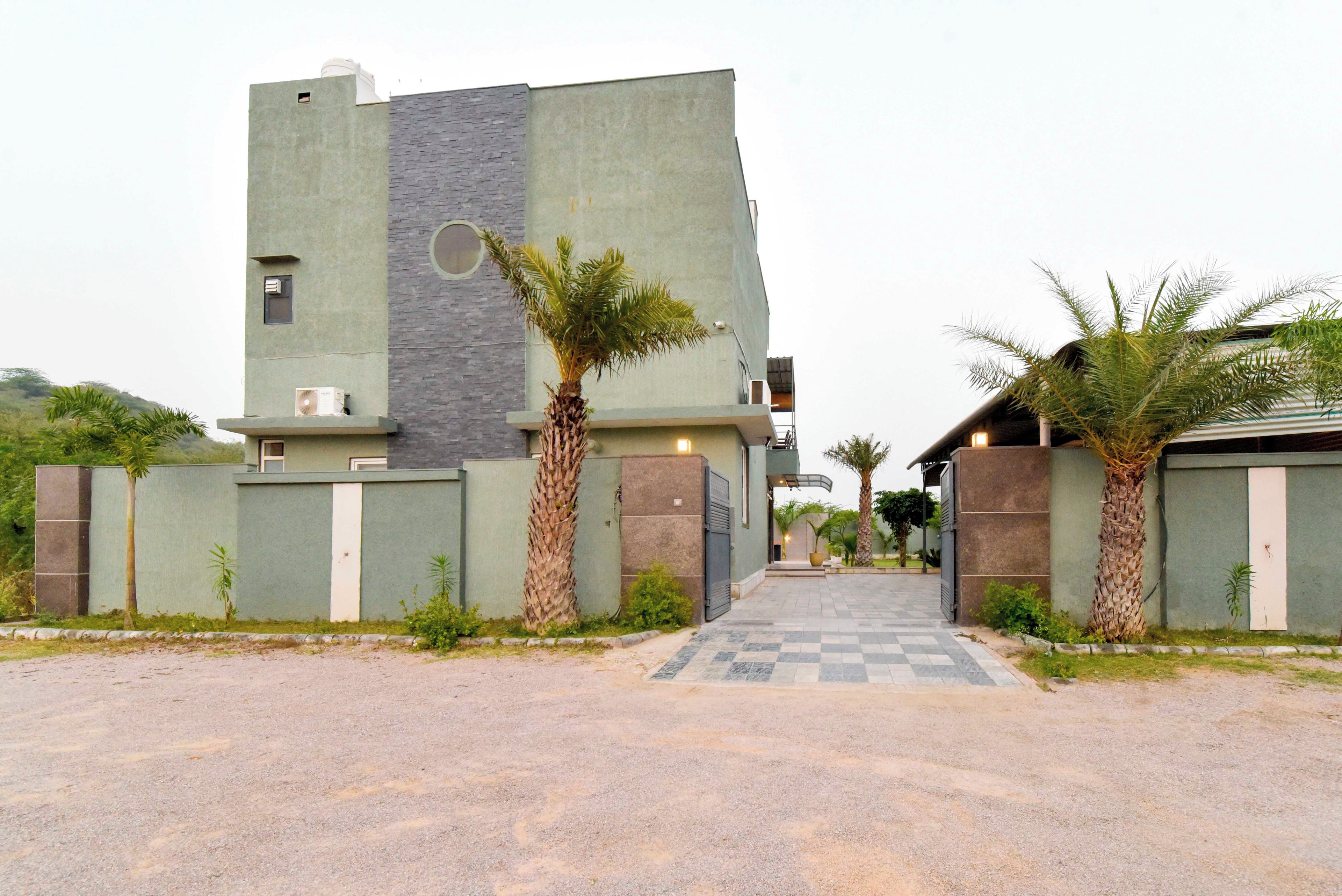 Un edificio moderno con un camino a cuadros que lleva a la entrada, rodeado de palmeras y una zona vallada.