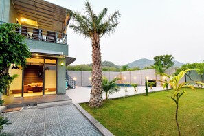 Un edificio moderno de dos pisos con entrada de vidrio, un área de estar al aire libre cubierta y un jardín ajardinado con una palmera.