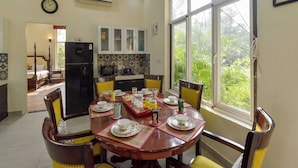 Uma área de refeições com uma mesa redonda posta para quatro pessoas, cercada por cadeiras com almofadas amarelas. Uma cozinha com armários e uma geladeira ao fundo.