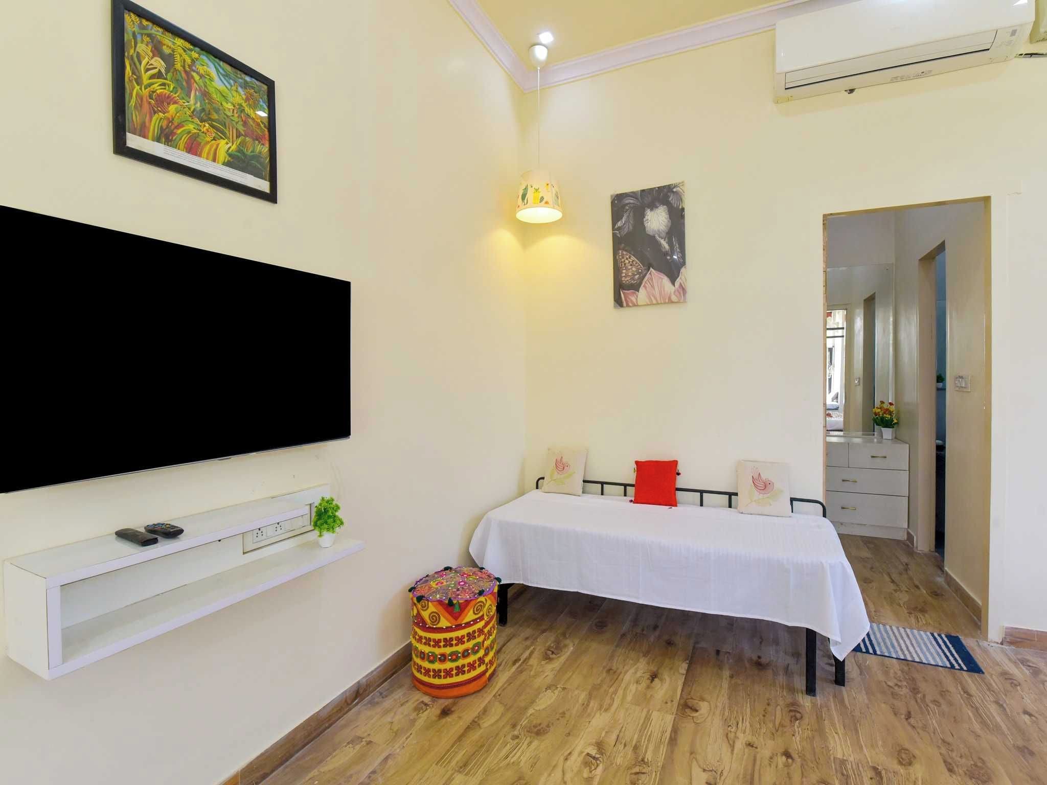 A room with a flat-screen TV mounted on the wall, a bed with a white bedspread, a small round table with a colorful top, and a wall-mounted light fixture.