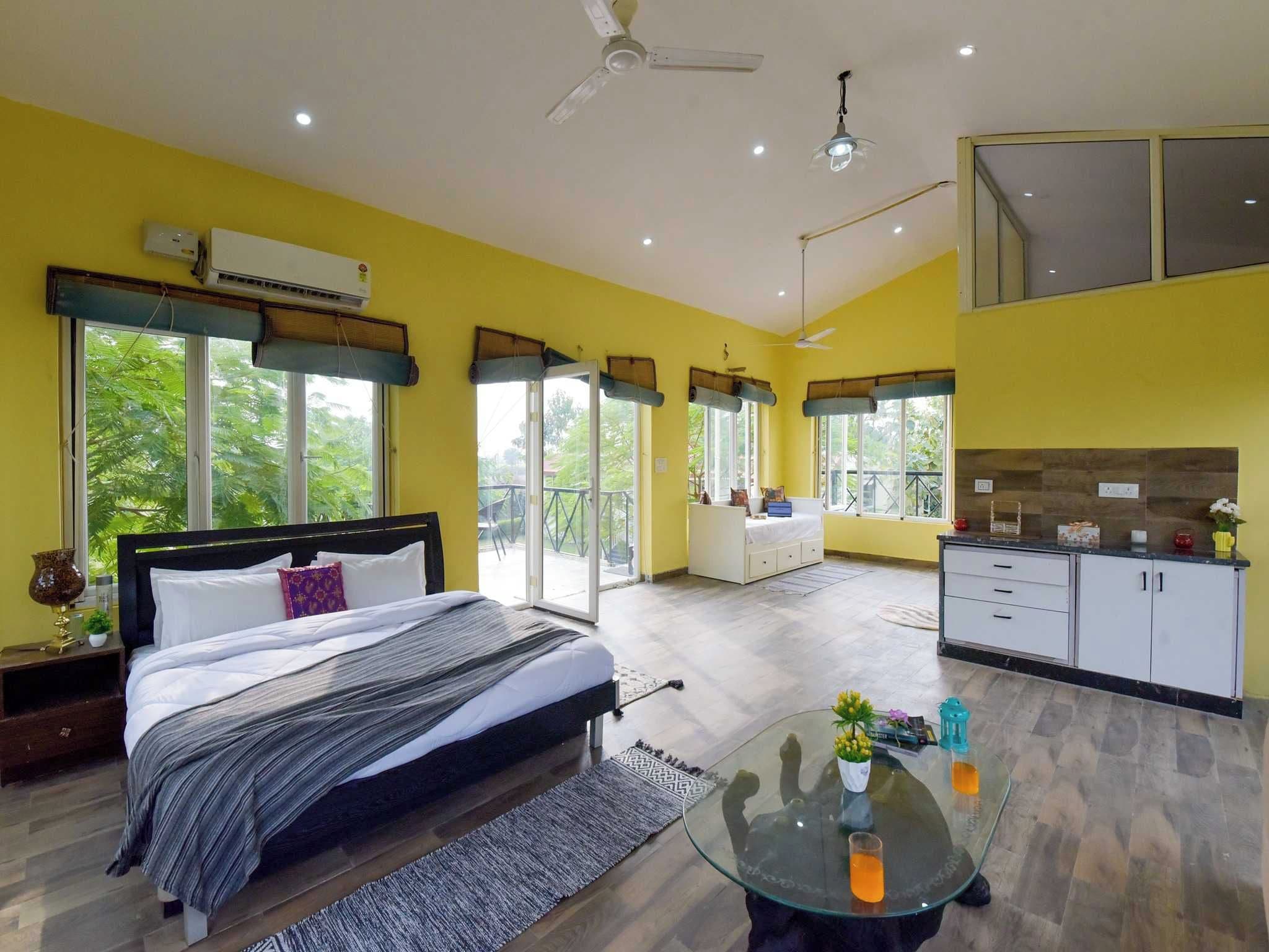A modern hotel room with a bed, a glass coffee table, a kitchen area, and large windows.