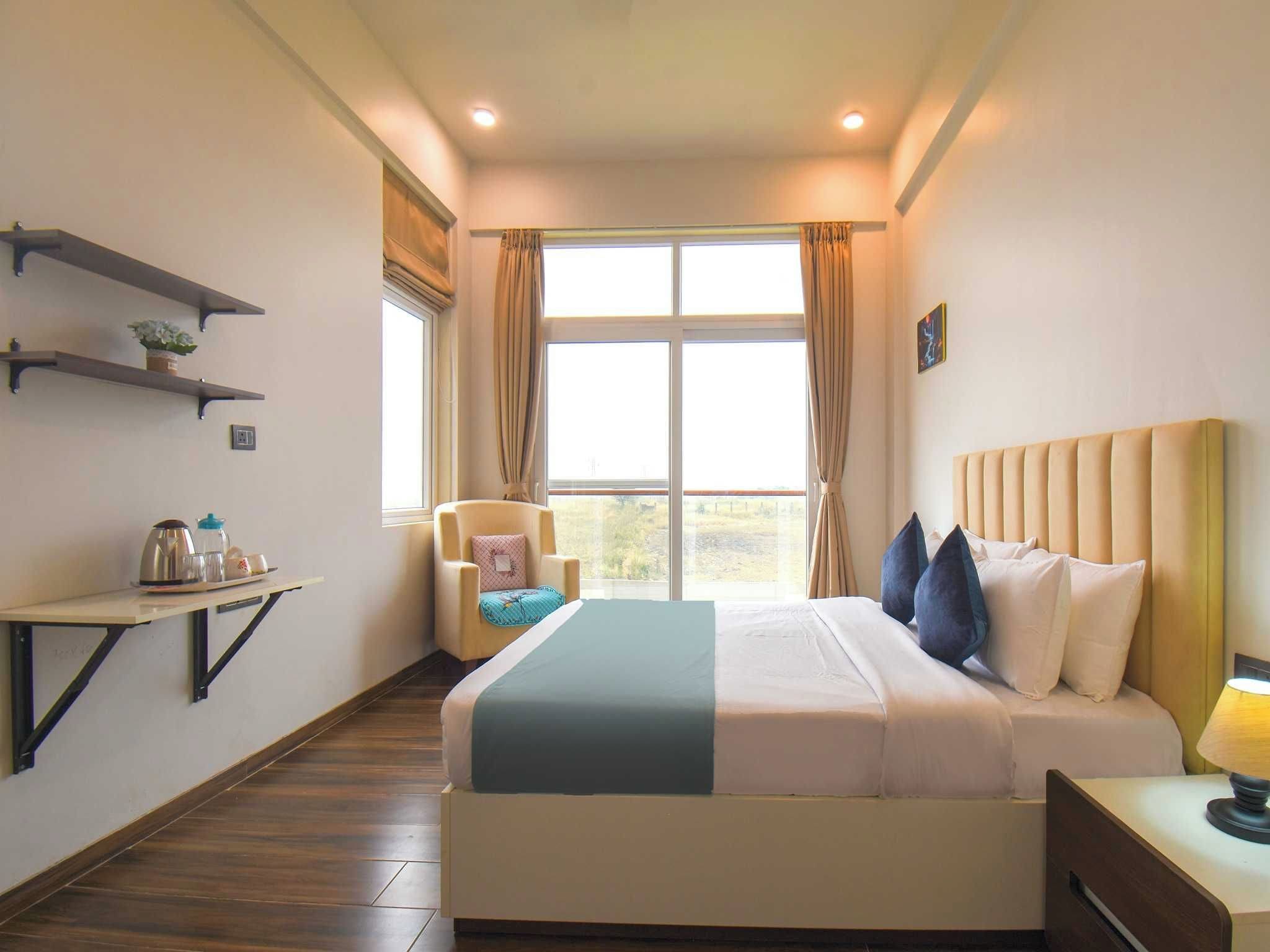 A hotel room with a bed, bedside table, chair, window, and a shelf with items.