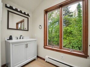 A bathroom with a white vanity, a mirror with lights, a window with a view of trees, and a wall-mounted heater. - Point Gratiot Lake House, Sleeps 12+, Lake Views (Dunkirk)