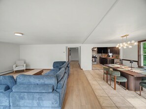 A modern living room with a long blue sofa, a small white armchair, and a dining area with a wooden table and chairs. - Point Gratiot Lake House, Sleeps 12+, Lake Views (Dunkirk)