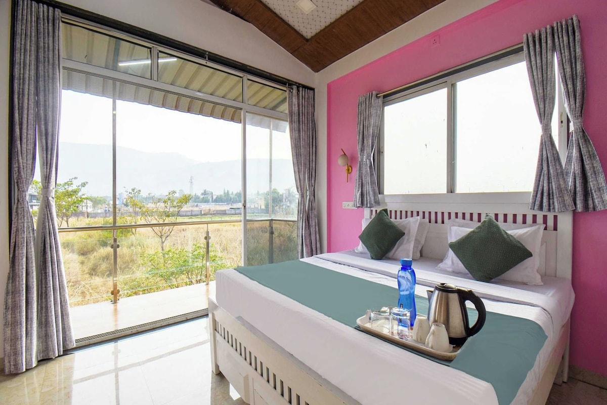 A hotel room with a large bed, a view of the countryside, and a pink wall.