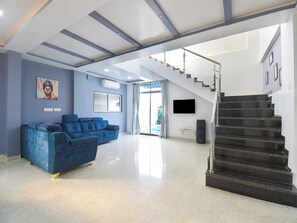 A spacious living area with a blue sofa set, a staircase, and a flat-screen TV mounted on the wall. - DanCenter Arohi Villa 4BHK Pvt Pool (Mawal)