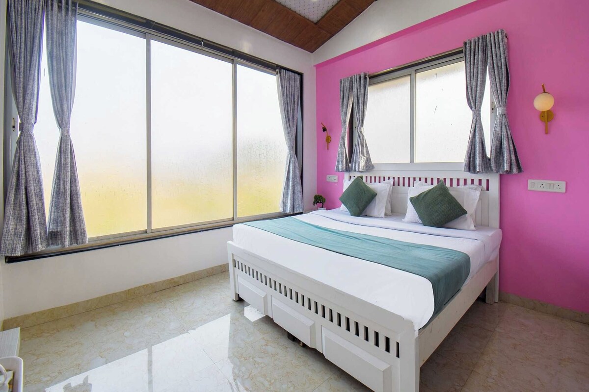 A bedroom with a large bed, two windows with curtains, a pink wall, and a ceiling with wooden beams.