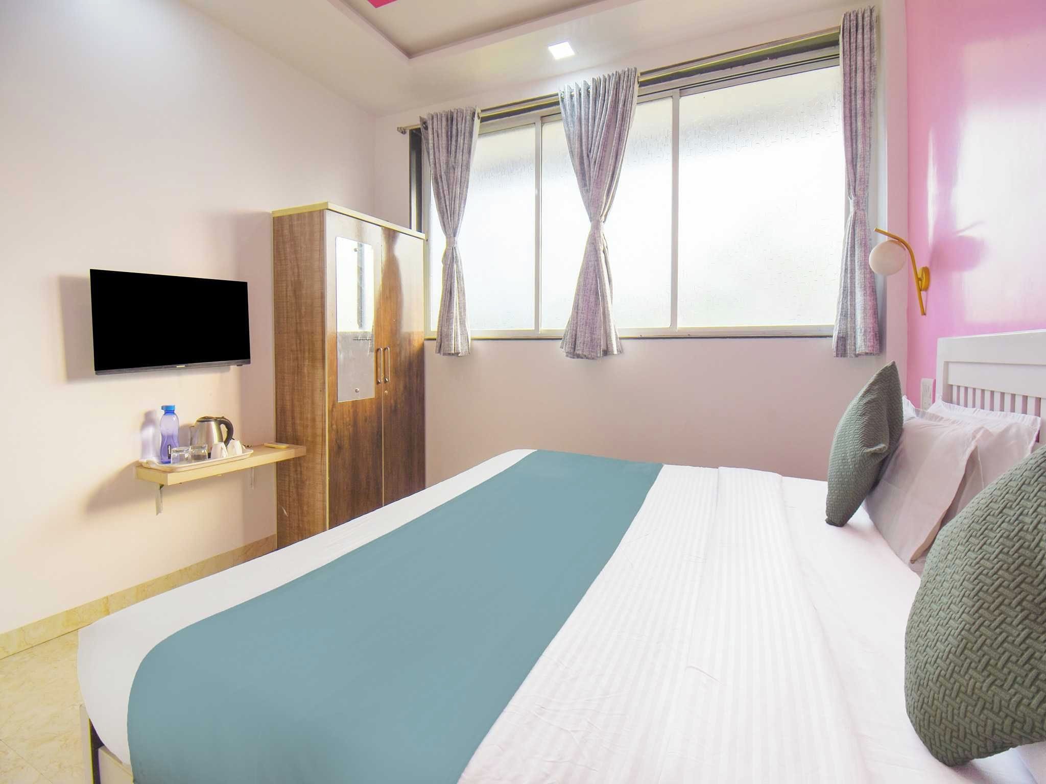 Una habitación de hotel con cama, televisor montado en la pared, armario y ventana con cortinas.