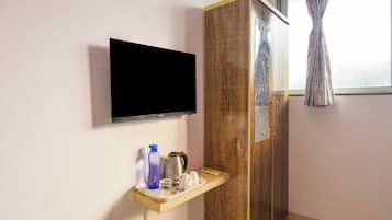 A room with a flat-screen TV mounted on the wall, a wooden wardrobe, and a small shelf with a kettle, cups, and bottles.