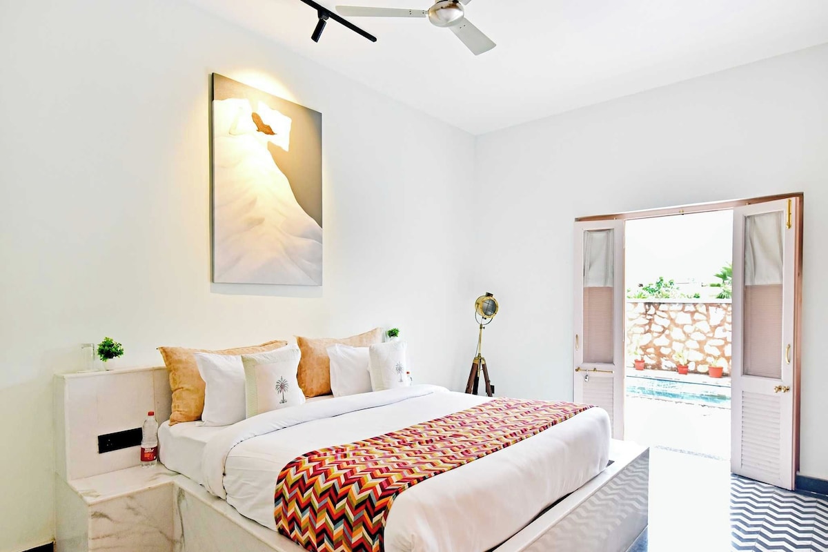 A hotel room with a bed, bedside tables, a wall-mounted lamp, a ceiling fan, and an open door leading to an outdoor area.