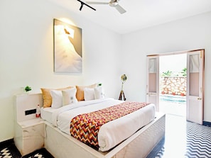 A hotel room with a bed, bedside tables, a wall-mounted lamp, a ceiling fan, and an open door leading to an outdoor area. - DanCenter Parijat Villas & Suites Pool (Udaipur)
