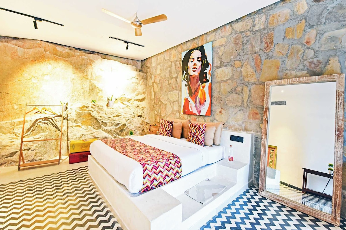 A bedroom with a stone wall, a bed with a patterned bedspread, a ceiling fan, and a large mirror.