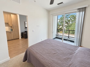 A bedroom with a large bed, a sliding glass door to a balcony, and a kitchen area visible through an open door.