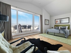Ein Schlafzimmer mit großem Fenster, einem Bett, einem Sessel und Blick auf den Balkon.