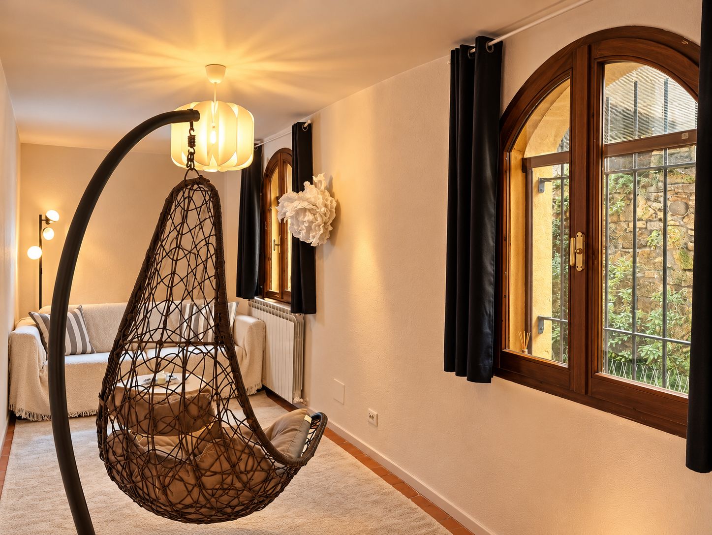 Une pièce avec un fauteuil suspendu, un radiateur, une fenêtre avec des rideaux et un luminaire fixé au mur.