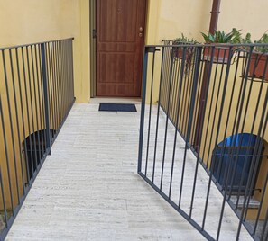 Un couloir étroit, carrelé, avec une porte en bois au bout, bordé de rambardes métalliques et de plantes en pot.