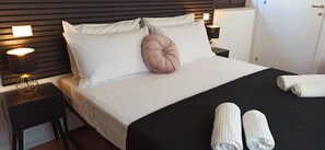 Ein ordentlich bezogenes Bett mit weißen Leinen, einem rosafarbenen Zierkissen und zwei aufgerollten Handtüchern.