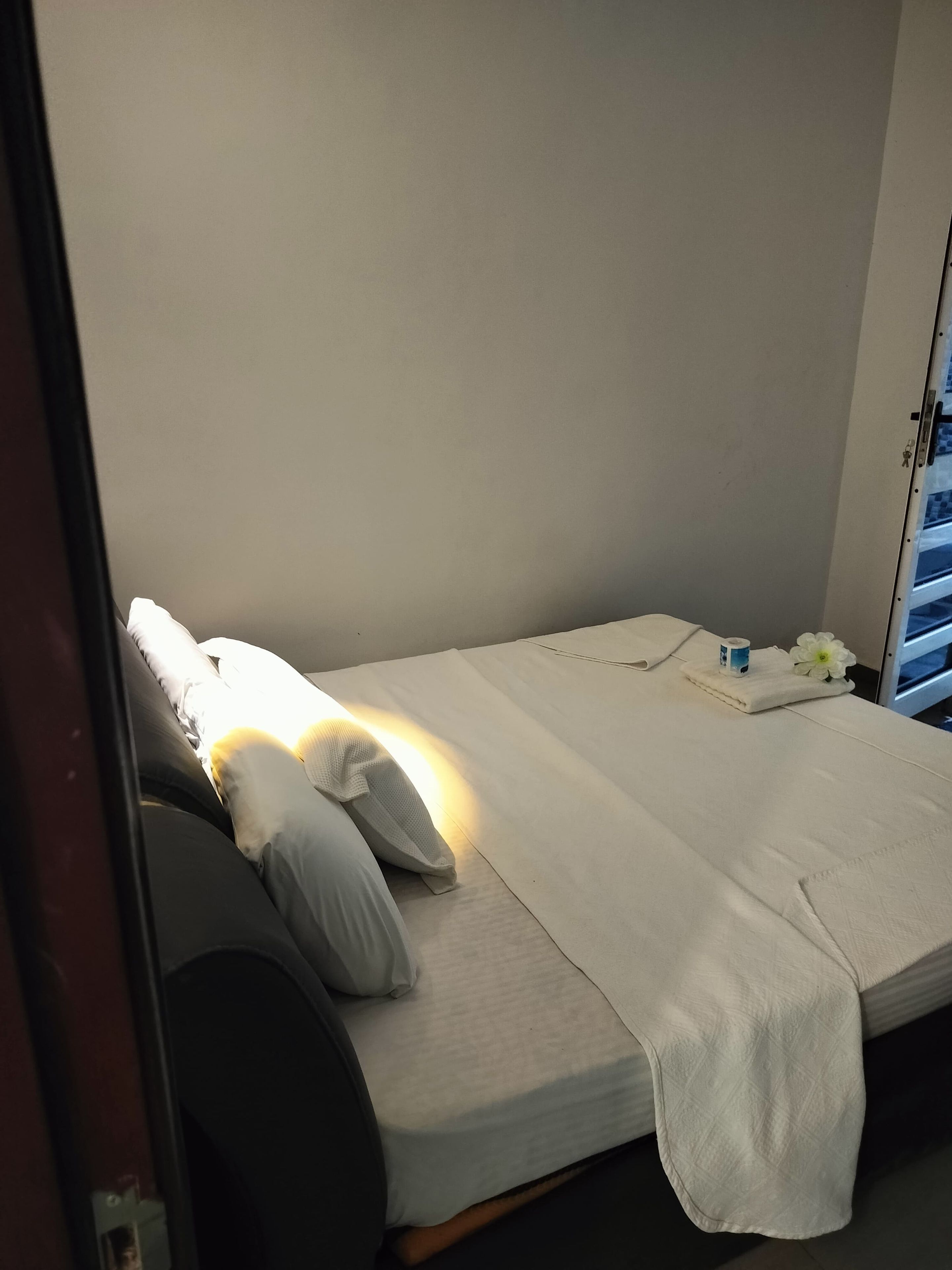 Una cama individual con ropa de cama y almohadas blancas, una mesita de noche con una caja de pañuelos y una flor, y una puerta con pomo.