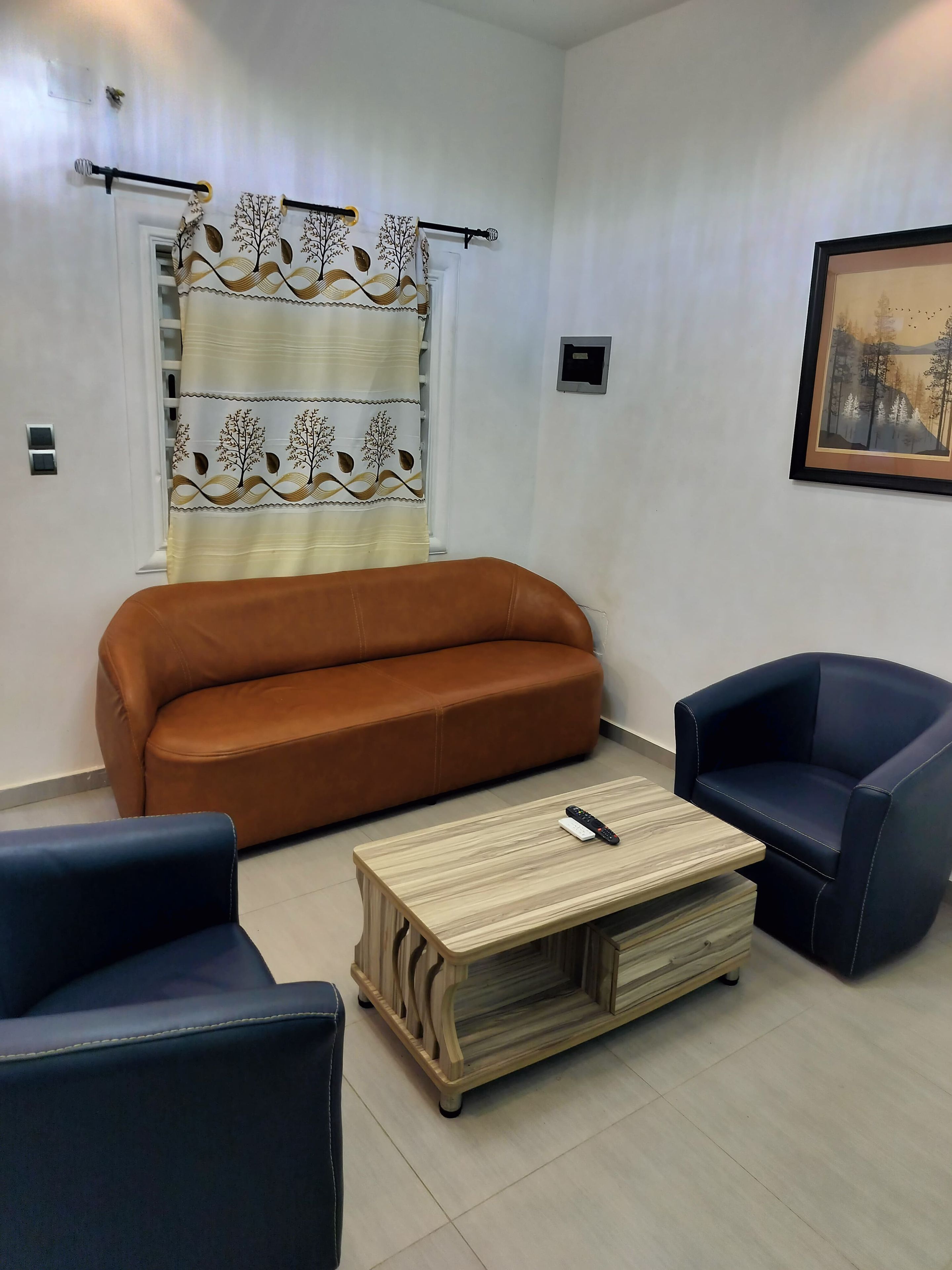 Ein Wohnzimmer mit einem braunen Sofa, zwei dunkelbraunen Sesseln, einem Holztisch und einem Vorhang mit Baum- und Blattmuster.