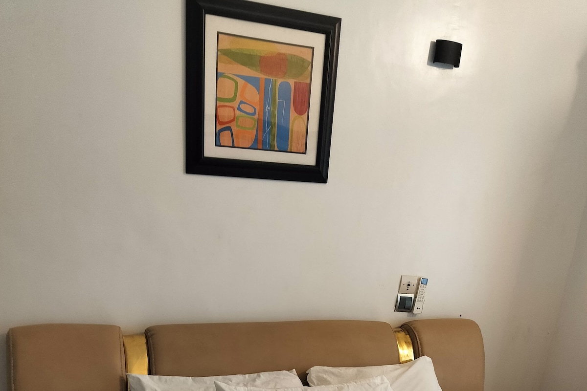 Een hotelkamer met een bed, kussens, een lijst met kunstwerk aan de muur en een bedieningspaneel aan de wand.