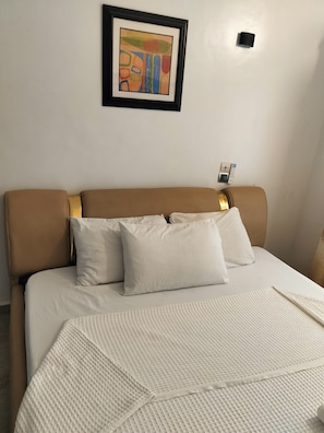 Een hotelkamer met een bed, kussens, een lijst met kunstwerk aan de muur en een bedieningspaneel aan de wand.