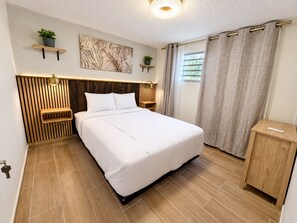 A modern bedroom with a wooden headboard, a bed with white bedding, a bedside table, a nightstand, and a wall-mounted shelf with a plant. - Gusto’s Inn_2 Queen Beds Sleeps 4 (Philipsburg)