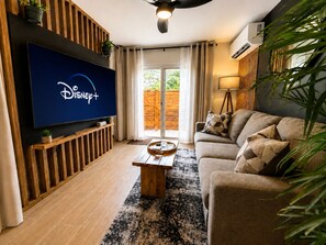 A living room with a flat-screen TV displaying the Disney+ logo, a wooden bench, a sofa with patterned cushions, and a large window with curtains. - Gusto’s Inn_2 Queen Beds Sleeps 4 (Philipsburg)