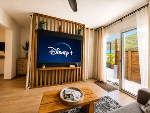 A living room with a flat-screen TV displaying the Disney+ logo, a wooden entertainment unit, a coffee table with remote controls, and a view of a hillside through large sliding glass doors. - Gusto’s Inn_2 Queen Beds Sleeps 4 (Philipsburg)