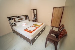 Un lit bien fait, avec un matelas blanc, une tête de lit en bois et une table de chevet marron sur laquelle se trouve une lampe. On y voit également une armoire en bois et une chaise en plastique marron avec un coussin.