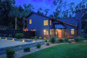 A two-story house with a dark siding exterior, a visible front porch with a bench, and a landscaped front yard with a stone retaining wall and various plants. - Loft of Legends-Vinyl Lounge/Walk to UF/Hot Tub/Sleeps 10 (Gainesville)