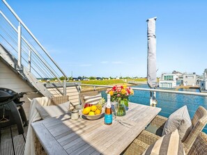 Eine Holzterrasse mit Korbmöbeln, einem Tisch mit Obstschale und Flasche, sowie Blick auf einen Swimmingpool und Gebäude im Hintergrund.
