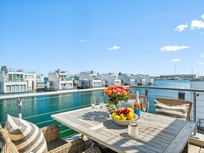 Eine Holzdielen-Terrasse mit Esstisch, mit Blick auf einen Yachthafen mit Booten und Gebäuden.