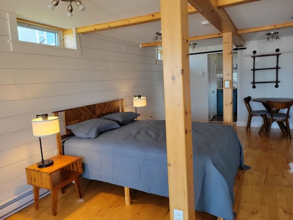 A bedroom with a bed, bedside lamps, a wooden headboard, and a dining area with a table and chairs. - The Sunset Harbour Village Suite (Chéticamp)
