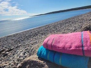 A rocky beach with a clear view of the water and a pile of colorful towels. - The Sunset Harbour Village Suite (Chéticamp)