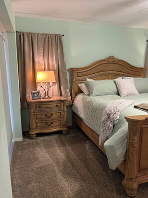 A bedroom with a wooden bed, a nightstand with a lamp, and a window with curtains. - Manatee Manor 3/2 House on Lake Sebring! (Sebring)