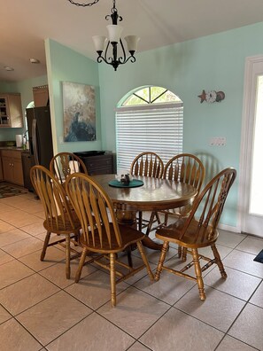 A dining area with a round table surrounded by wooden chairs, a chandelier, and a window with blinds. - Manatee Manor 3/2 House on Lake Sebring! (Sebring)